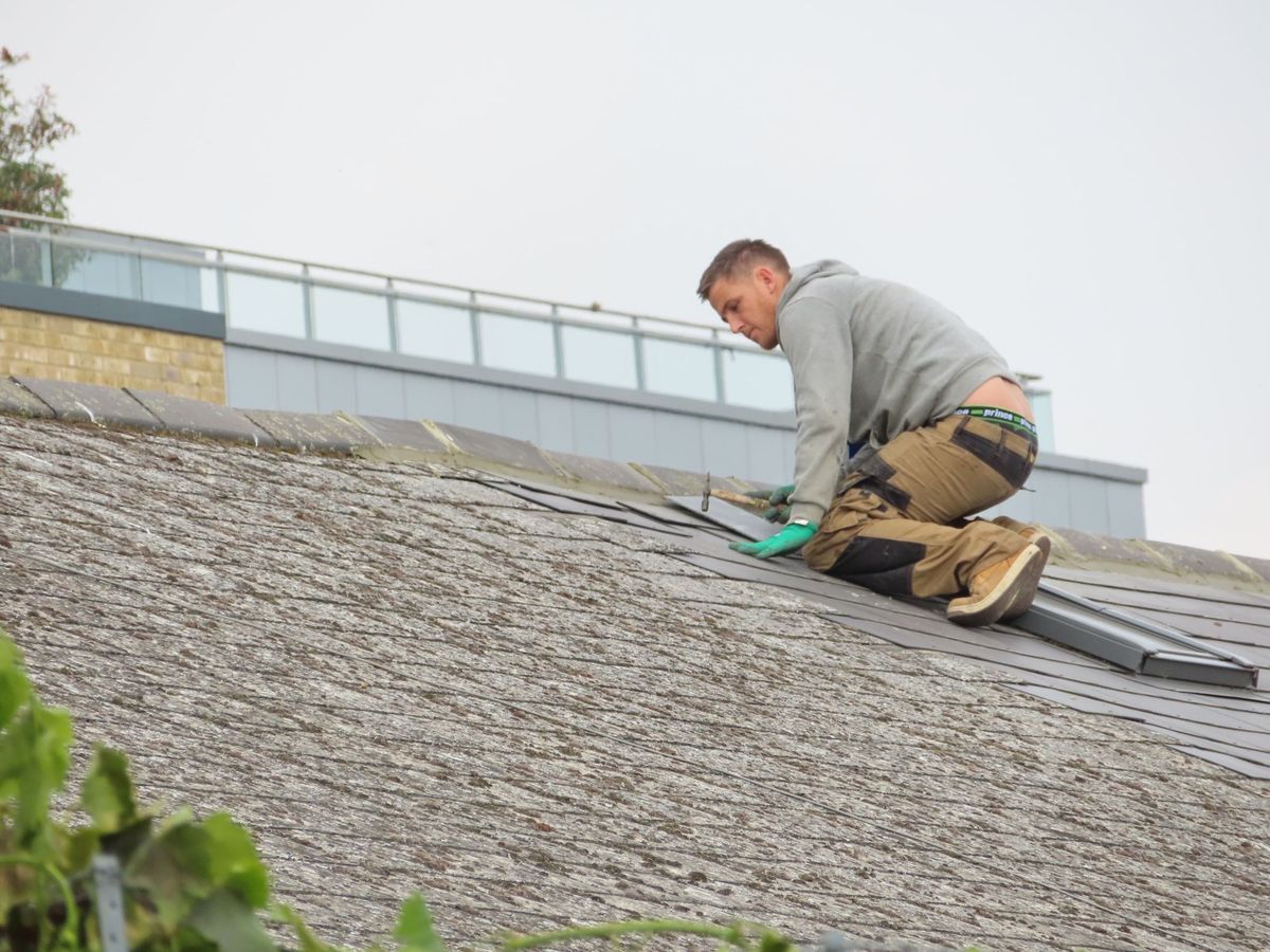 A roofer kneeling on a pitched roof checking the shingles